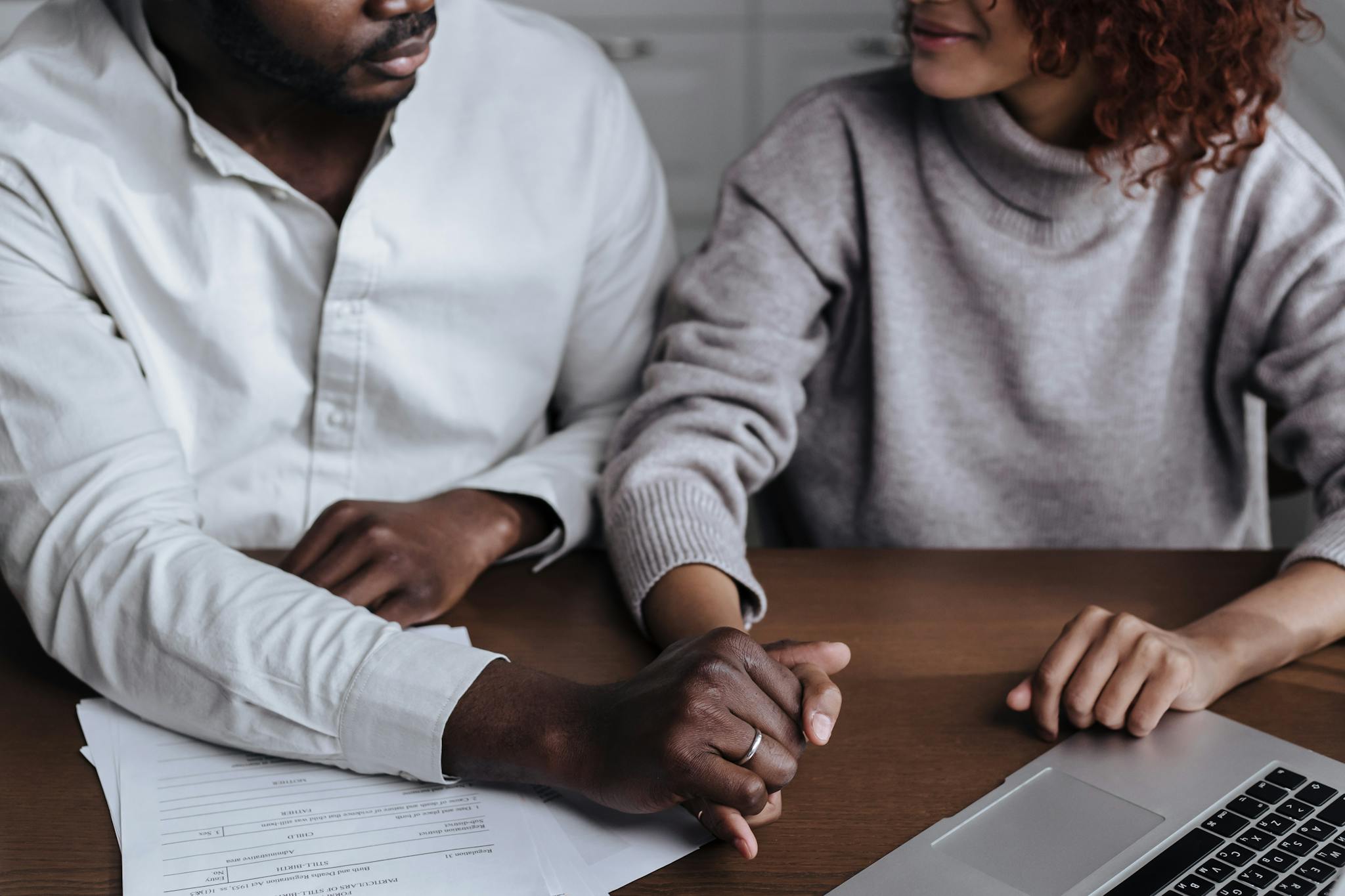 A diverse couple holding hands with a laptop and paperwork on the table.