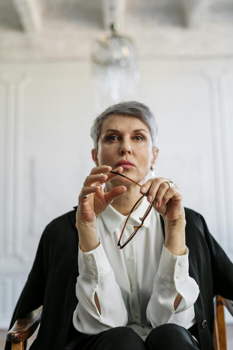 A mature woman in deep thought holds her glasses in a serene, stylish interior setting.