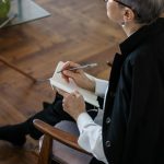 A professional woman with glasses taking notes while seated indoors, depicting consultation or therapy.