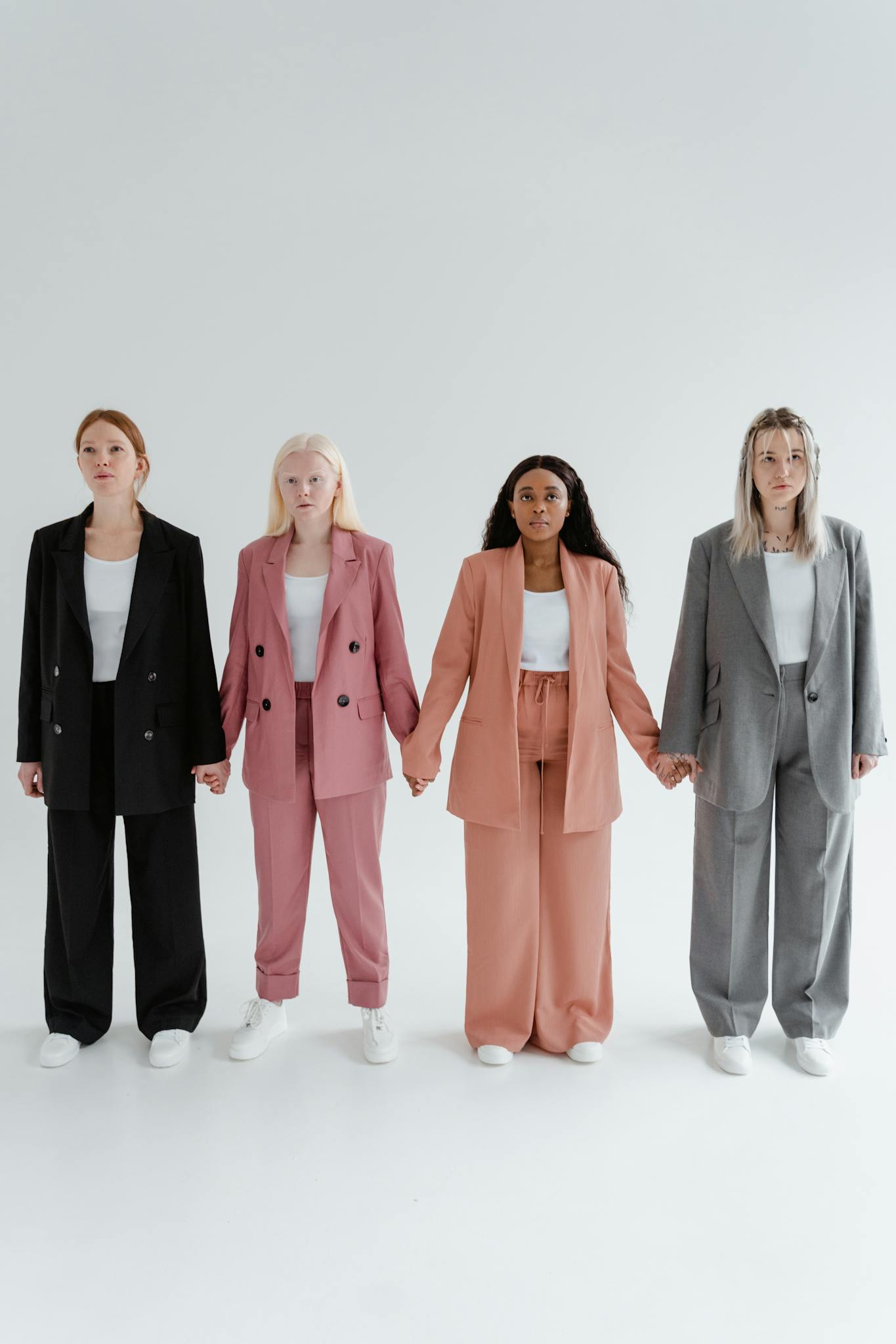Four diverse women in stylish business suits holding hands on a white background.