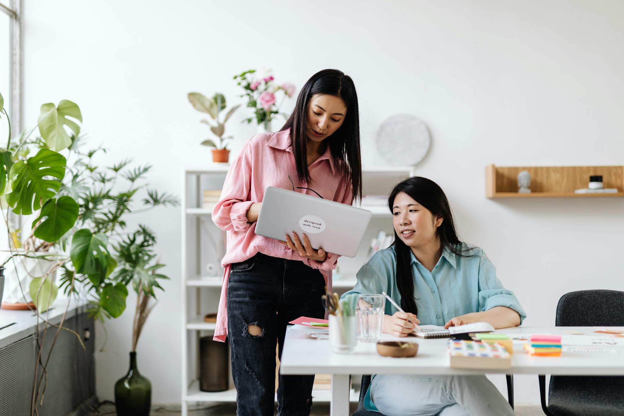 Two women engaged in teamwork using a laptop, surrounded by office plants and supplies.